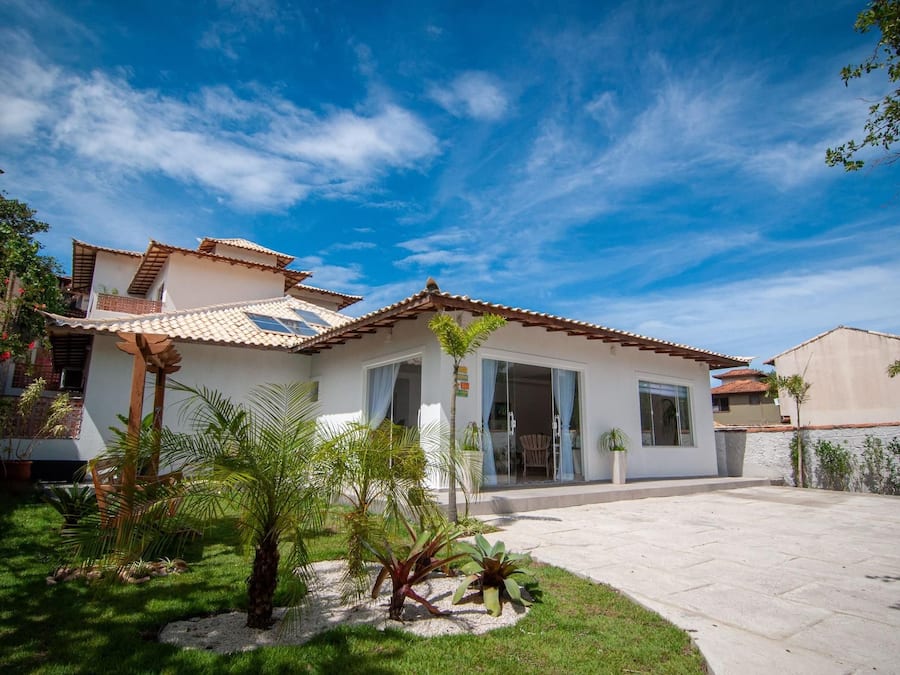 Uma casa de dois andares com telhado de telhas, varanda frontal e um jardim bem cuidado, com palmeiras e arbustos.