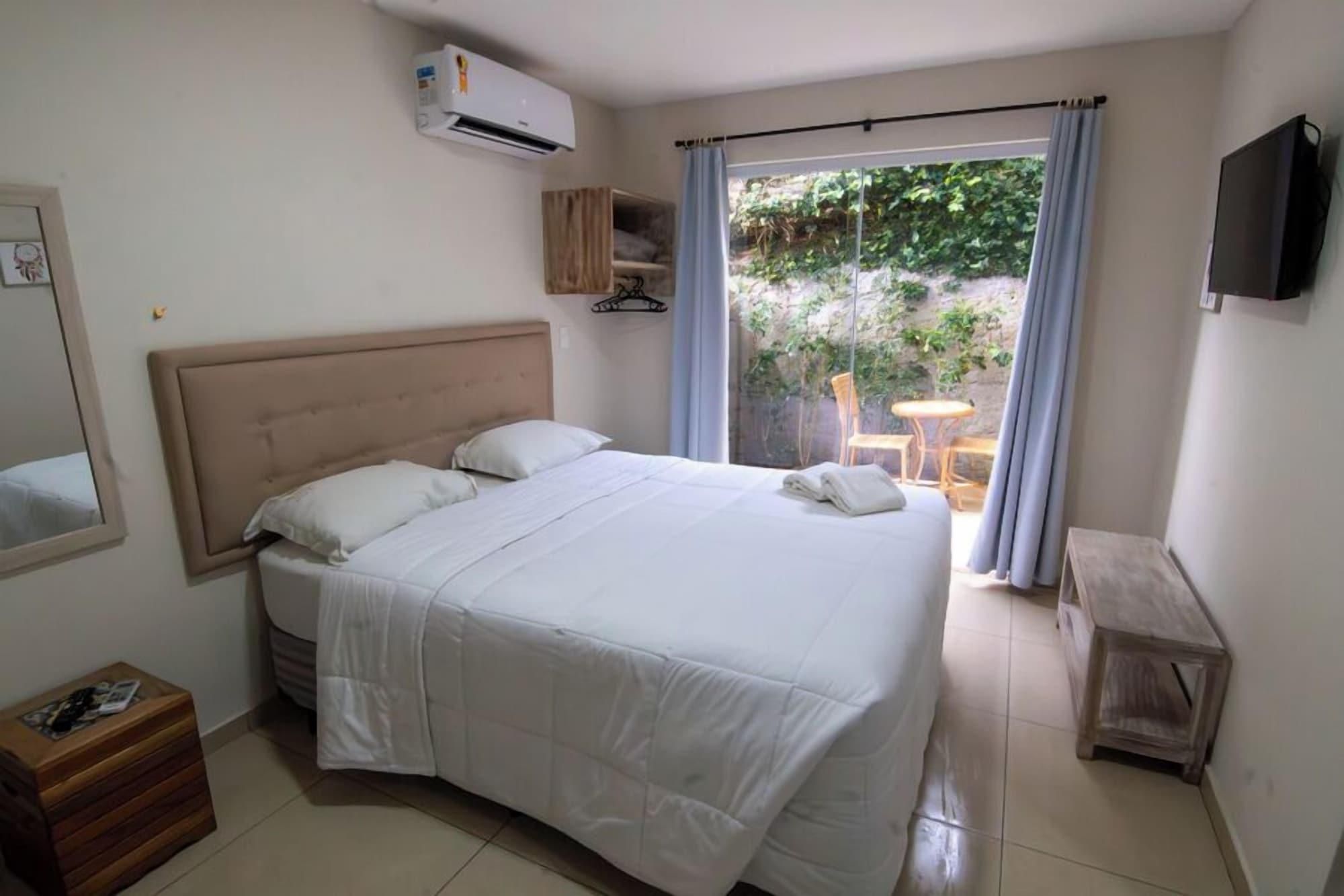 Quarto com cama, um baú de madeira, ar condicionado, TV e vista para área externa com mesa e cadeiras.