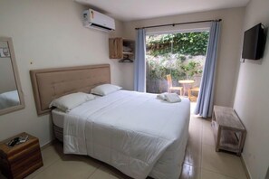 A bedroom with a bed, a wooden chest, an air conditioner, a TV, and a view of an outdoor area with a table and chairs. - Pousada Maktub (Búzios)