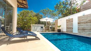 Una piscina con sillas de descanso y una mesa, rodeada por un muro blanco con paneles de madera y un ventanal.
