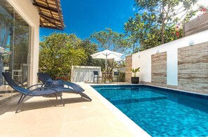 Una piscina con sillas de descanso y una mesa, rodeada por un muro blanco con paneles de madera y un ventanal.
