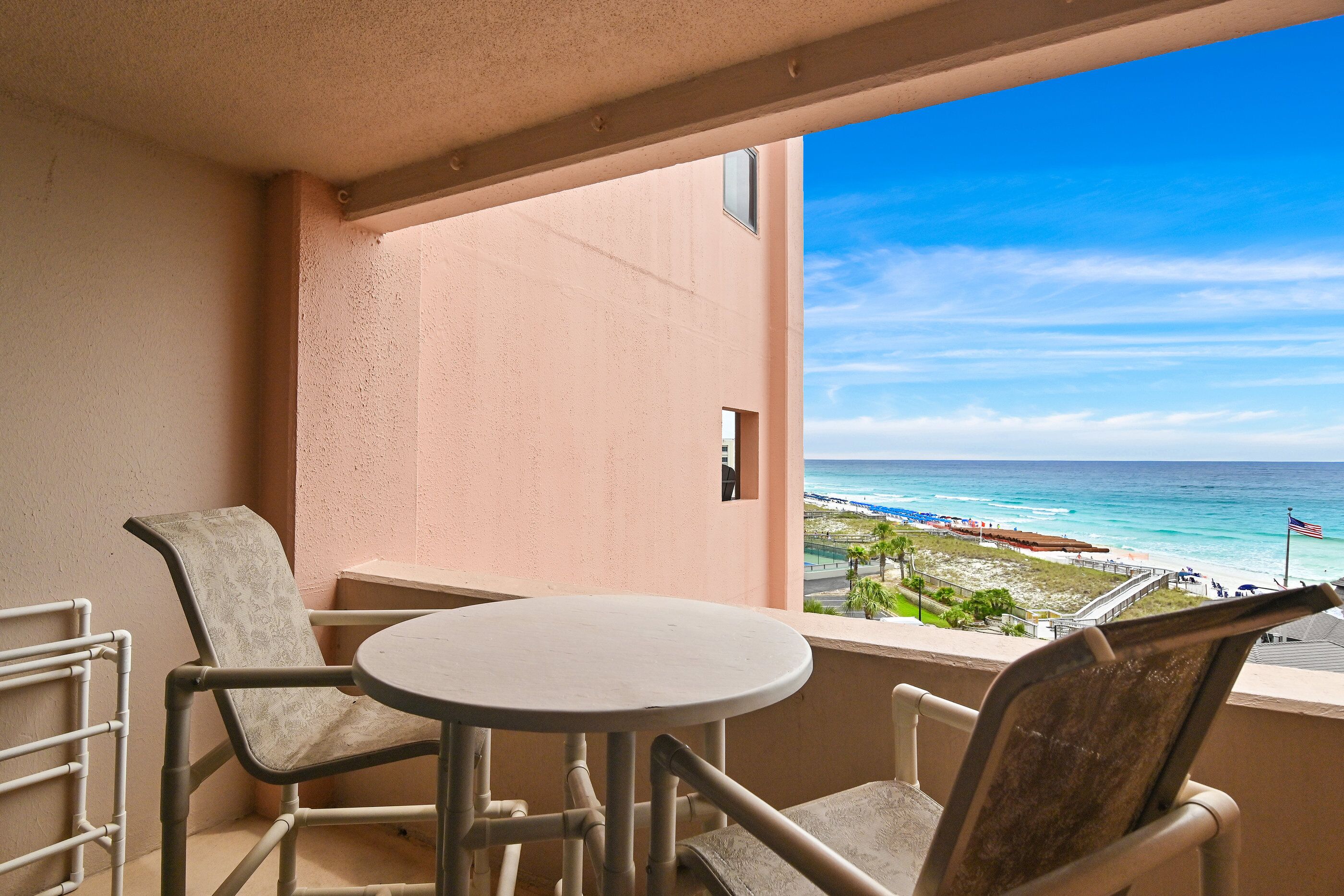 A balcony with a round table and two chairs, overlooking a beach with a clear sky.