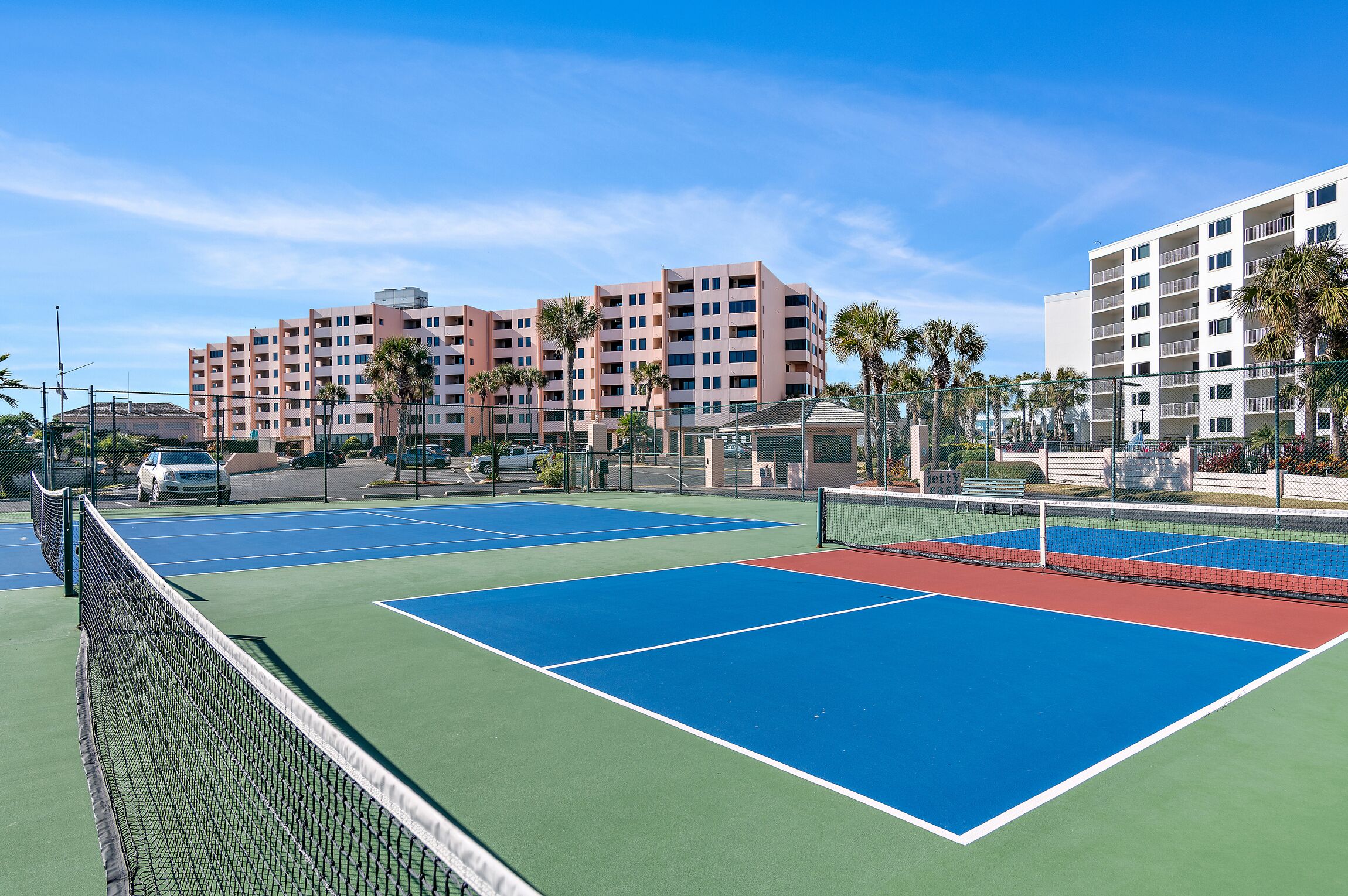 Un court de tennis avec un filet, entouré d’une clôture, et situé à côté d’un immeuble à plusieurs étages doté de balcons et de palmiers.