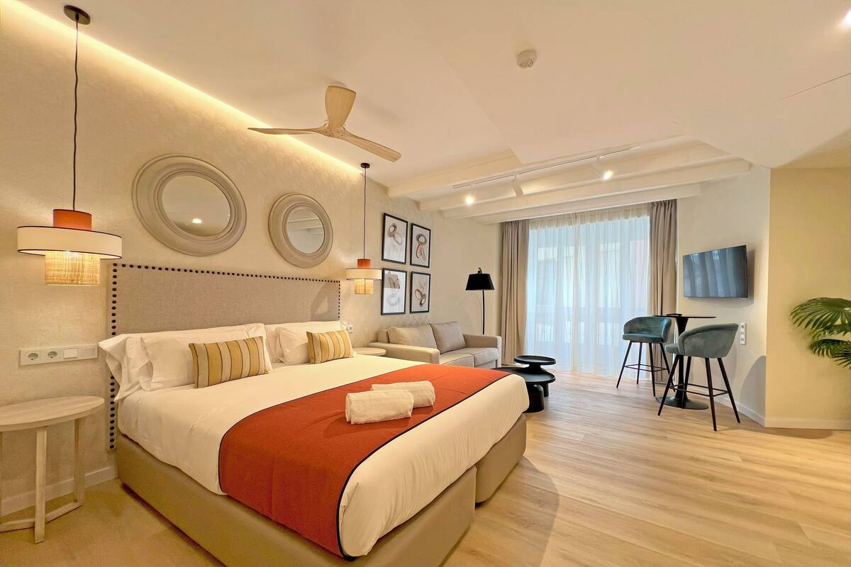A modern hotel room with a large bed, a ceiling fan, a TV, a seating area with a sofa and chairs, and a large window with curtains.