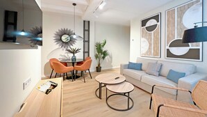 A modern living room with a round dining table, orange chairs, a grey sofa, and abstract wall art. - Apartamentos Soho Boutique Palillero (Cádiz)