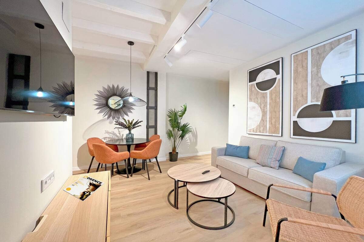 A modern living room with a round dining table, orange chairs, a grey sofa, and a large abstract painting on the wall.