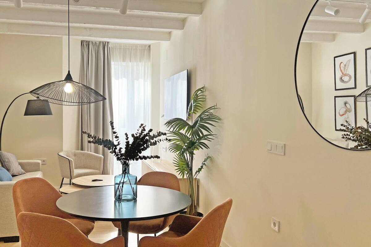 A modern living room with a round dining table, orange chairs, a sofa, a coffee table, and a large window with curtains.