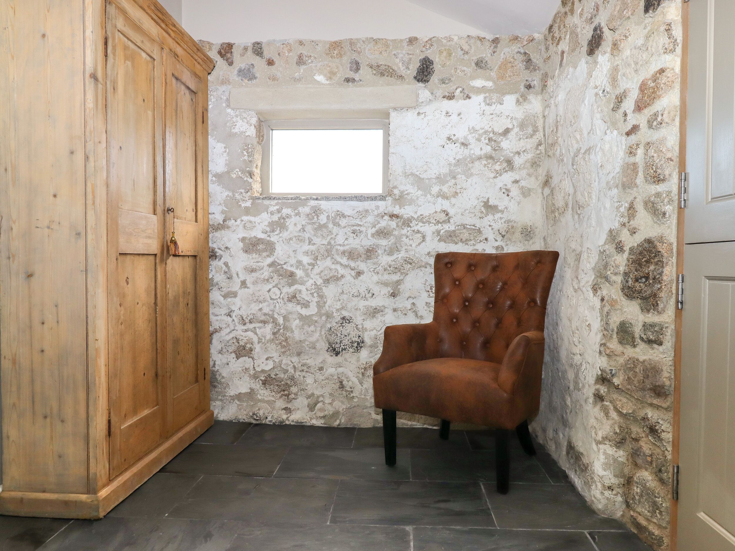 A room with a stone wall, a wooden wardrobe, a window, and a brown chair.