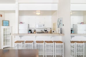 A modern kitchen with white cabinets, a microwave, and a coffee machine. There are four stools with woven seats and a wooden table in the foreground. - Sand Dollar Beachfront Home, Direct Beach Access! (La Selva Beach)