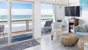 Une pièce spacieuse dotée d’une grande porte-fenêtre coulissante donnant sur un balcon avec vue sur l’océan. Un téléviseur à écran plat est fixé au mur, et un coin salon confortable est aménagé avec un canapé et un pouf.