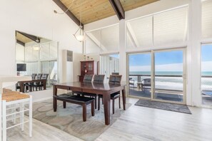 Una habitación amplia con una gran mesa de madera, sillas y vista al océano a través de amplios ventanales correderas.