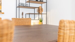 A dining area with a dark wooden table, wicker chairs, a black metal shelving unit with decorative items, and a potted plant. - Apartment "Avenida Masnou" close to the Sea with Shared Pool & Wi-Fi (Calpe)