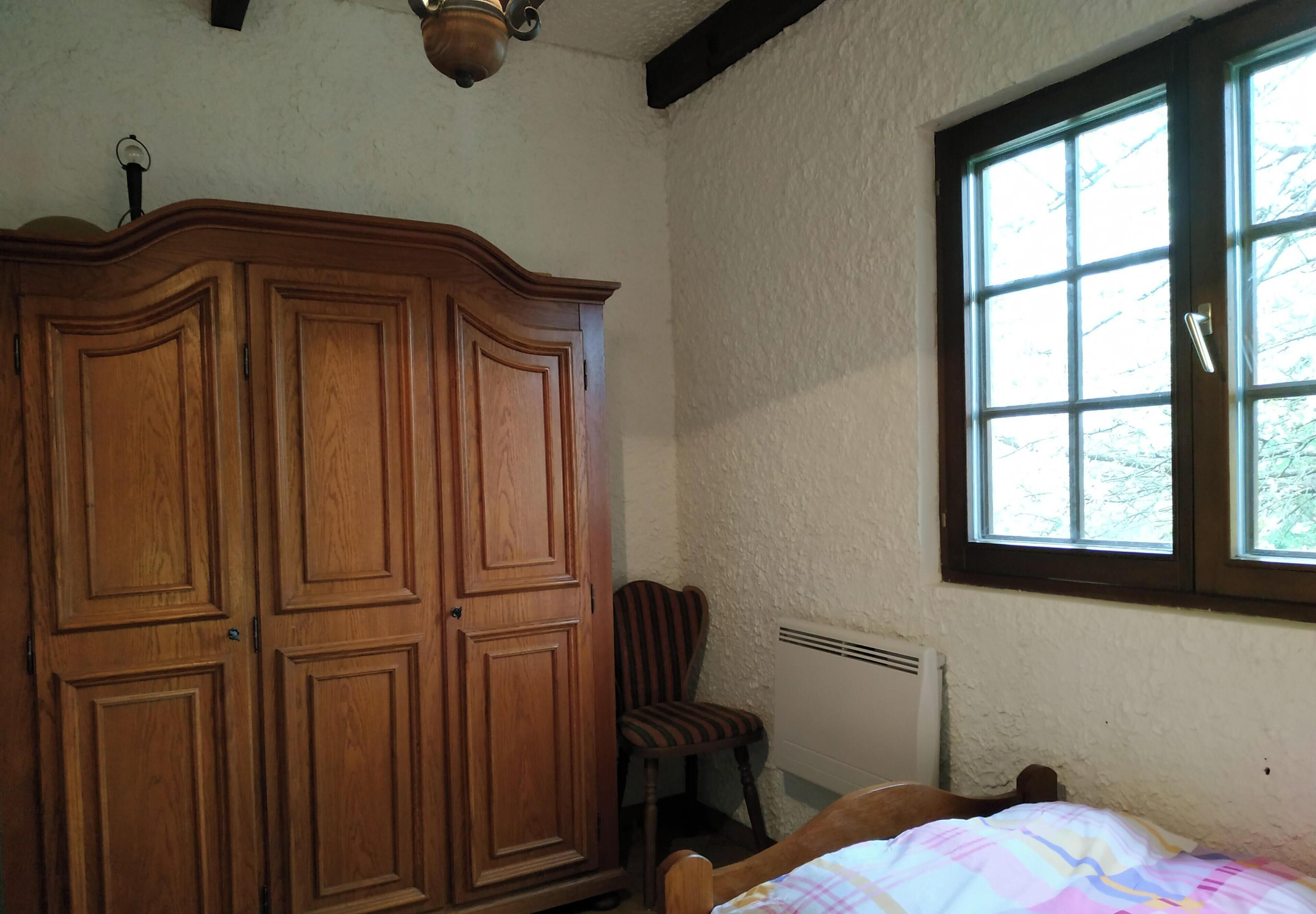 Una camera da letto con un'ampia armadio in legno, una sedia singola, una finestra vista alberi e un termosifone.