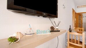 A flat-screen TV mounted on a wall, a wooden shelf with decorative items, a table with a patterned tablecloth, and a chair. - Vacation Home "Las Arenas Bajo" close to the Sea with Wi-Fi (Las Arenas)