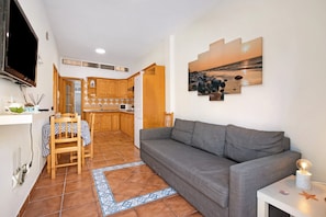 Sala de estar com sofá cinzento, mesa de jantar com cadeiras, cozinha ao fundo e televisão na parede.