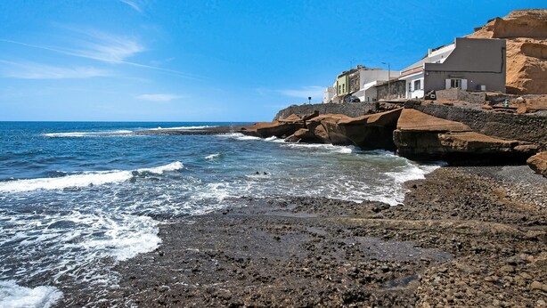 Escena costera con terreno rocoso, una playa y edificios de estilo arquitectónico mediterráneo.