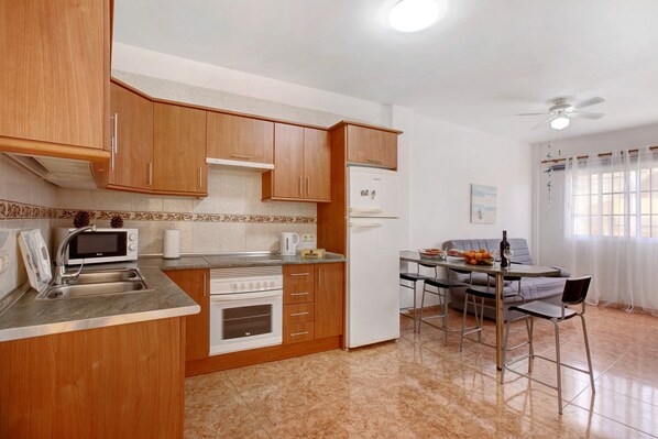 A compact kitchen with wooden cabinets, a microwave, a refrigerator, a dining table with chairs, and a ceiling fan.
