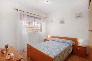A bedroom with a wooden bed, a nightstand with a lamp, a small table with a clock and decorative items, and a window with sheer curtains.