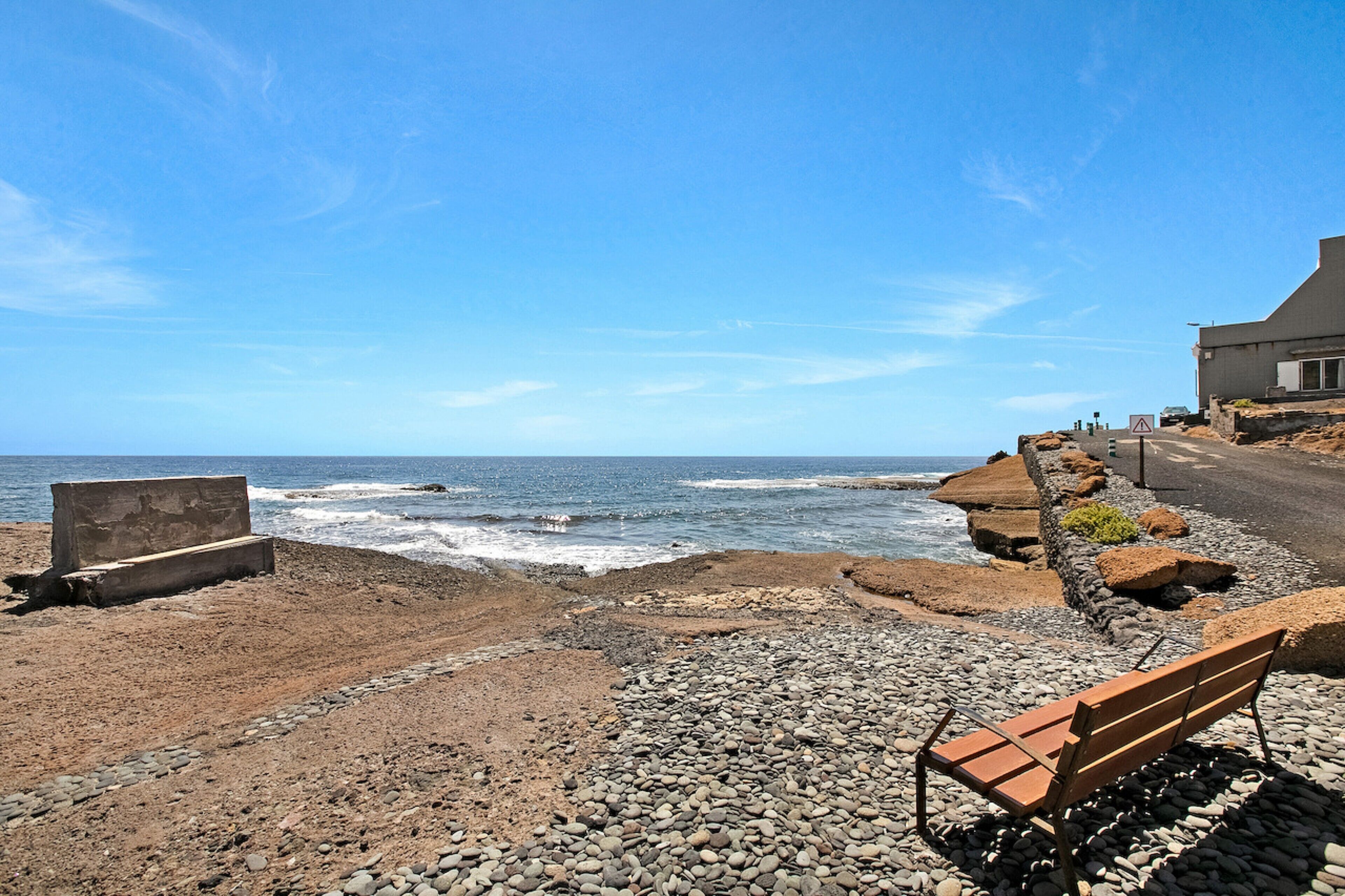 Una playa rocosa con un banco, un bloque de concreto y un edificio al fondo.