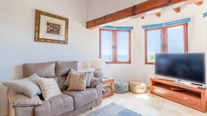 Una sala de estar con sofá, televisor, vigas de madera, ventanas y un cuadro enmarcado en la pared.