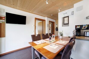 Un espace repas comprenant une table en bois, des chaises, un téléviseur et un coin cuisine équipé d’une machine à café.