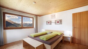 Ein Schlafzimmer mit Holzbalkendecke, einem Bett mit grün-violetter Bettwäsche, einer Holzbank, einem Nachttisch, einer Nachttischlampe und Bergblick durch ein Fenster.