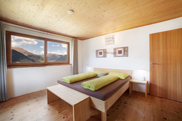 Une chambre avec un plafond en bois, un lit avec une literie verte et violette, un banc en bois, une table de chevet, une lampe de chevet et une vue sur les montagnes par la fenêtre.