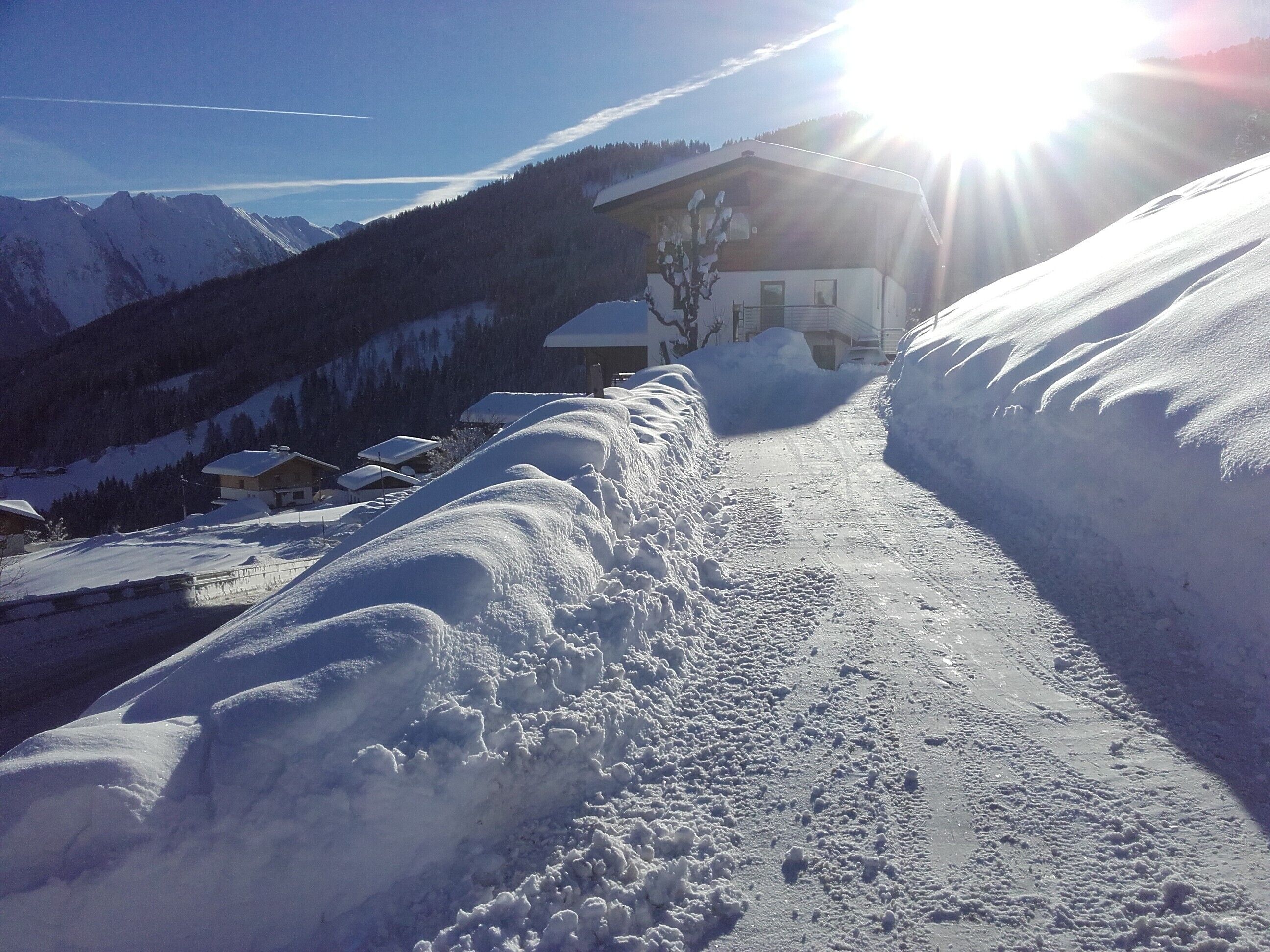 Ein verschneiter Bergpfad führt zu einem Gebäude mit schneebedecktem Dach, umgeben von Schneewehen und einem klaren Himmel.