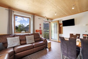 A modern living room with a brown leather sofa, a dining table with chairs, a flat-screen TV, and a large window with a mountain view. - Apartment 'Joshof Bergblick' with Mountain View, Balcony and Wi-Fi (Moso in Passiria)