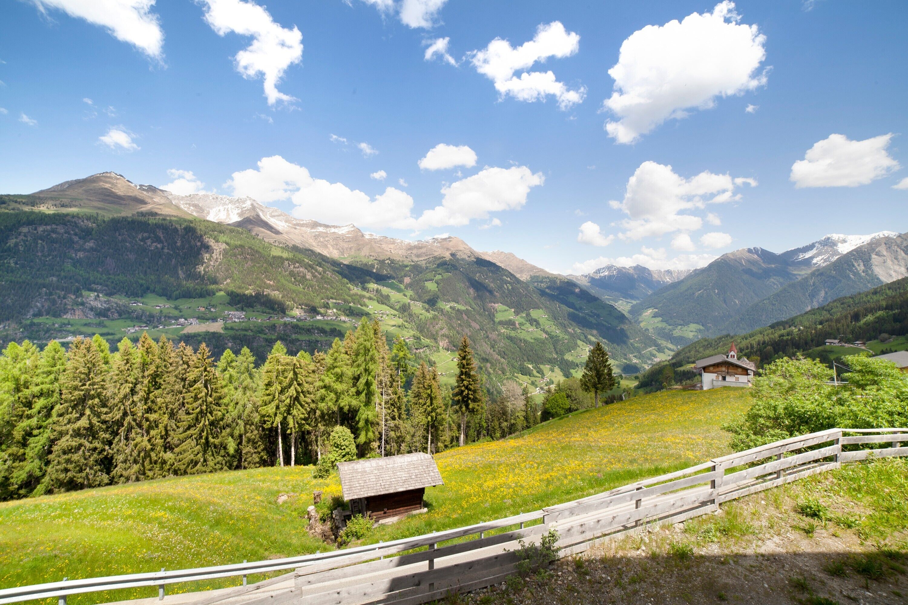 A mountainous landscape with a wooden fence, a small cabin, and distant mountains under a clear sky.