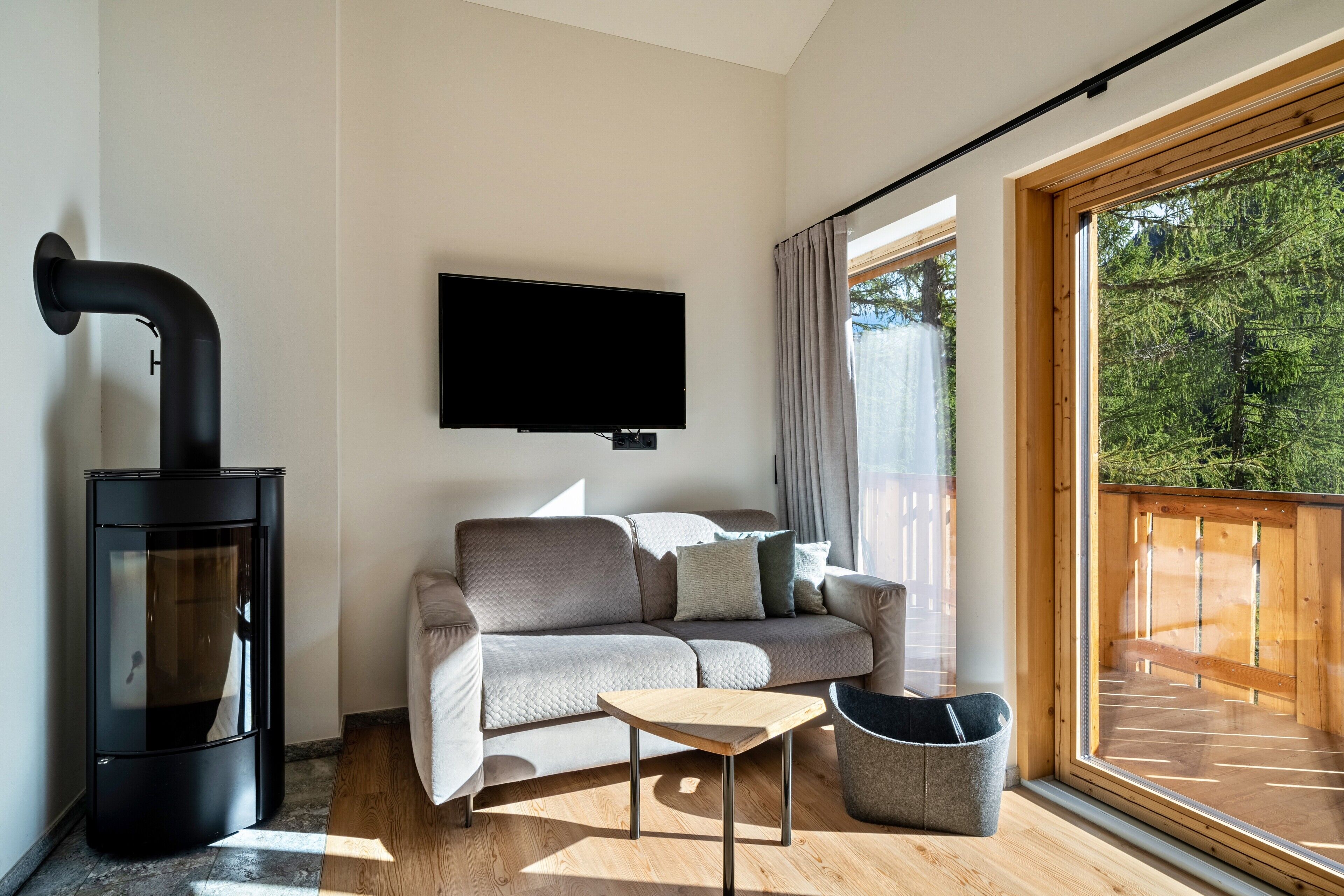 En moderne stue med sofa, et lille bord, en sort brændeovn, et tv ophængt på væggen og et stort vindue med udsigt til træer.