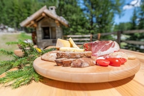 Una tabla de madera con varios quesos, embutidos y tomates sobre una mesa, con una casa rústica y follaje en el fondo.