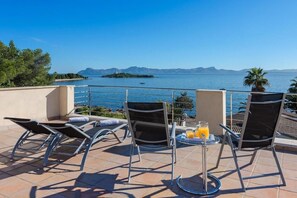 A terrace with lounge chairs, a table with a drink, and a view of the sea and mountains.