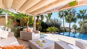 A covered outdoor seating area with wicker furniture, a pool, and tropical plants.