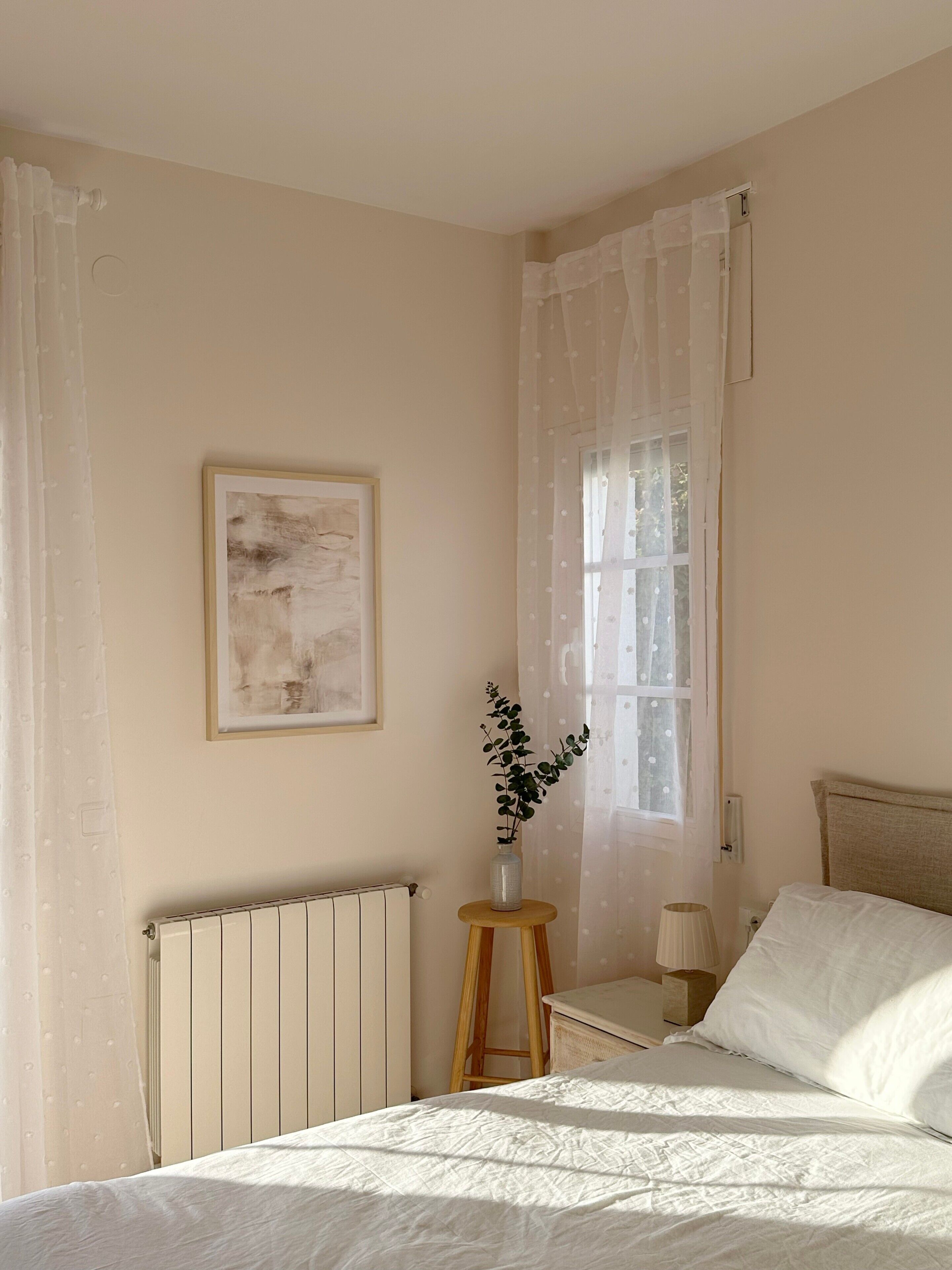 Et soveværelse med seng, radiator, et vindue med let gardin, et lille bord med lampe og et billede i ramme på væggen.