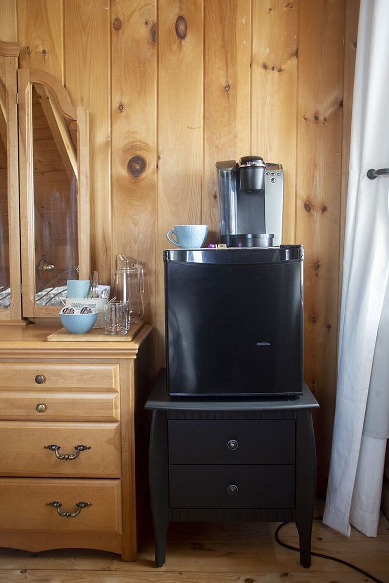 A wooden cabin interior with a black refrigerator, a wooden dresser with drawers, and a mirror.