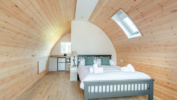 Un dormitorio con techo curvo, tragaluz, cama con almohadas y toallas, mesita de noche y pequeño escritorio con lámpara.