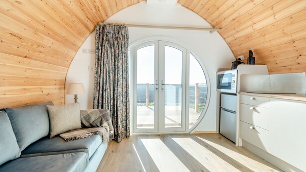 Een gezellig chalet binnen met een bank, gordijnen, een keukenhoek en een grote glazen deur die uitkomt op een terras.