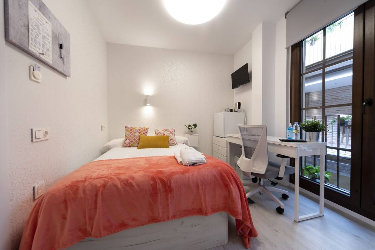 A modern bedroom with a bed, desk, and large window.