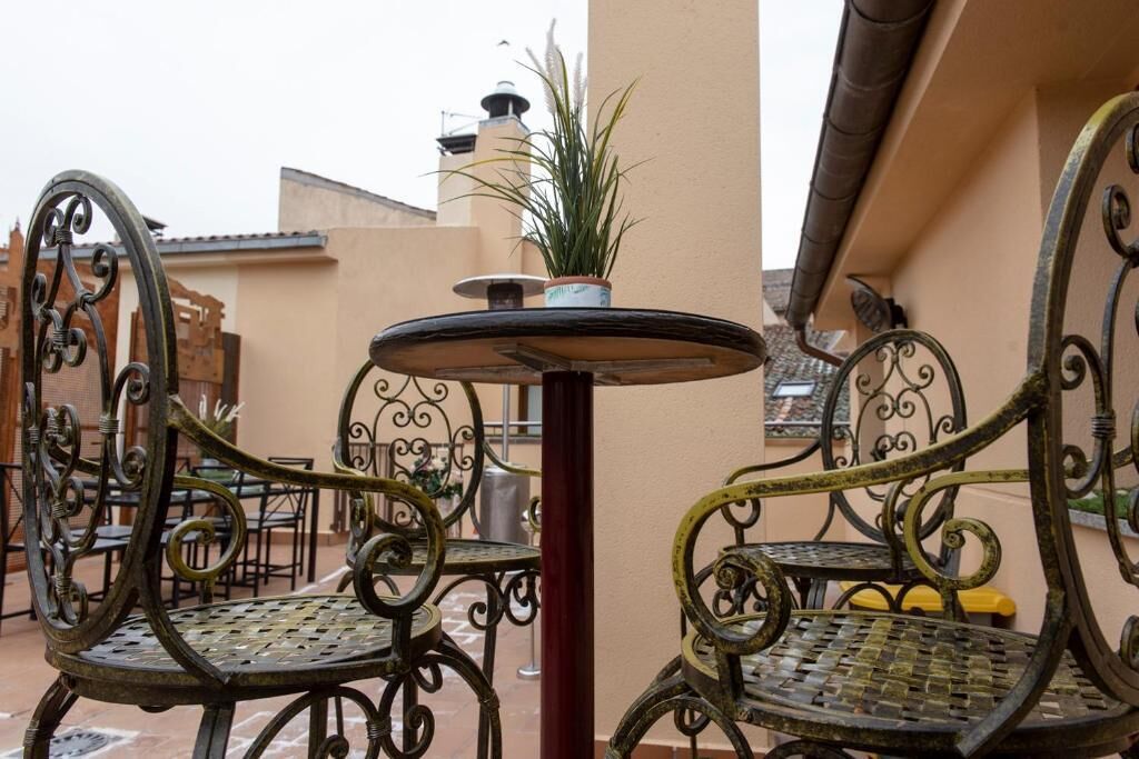 An outdoor patio area with wrought iron chairs and a small round table, surrounded by beige walls and a tiled roof.