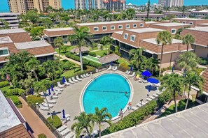 A resort with a central swimming pool surrounded by lounge chairs and umbrellas, palm trees, and a view of the city skyline. - Quaint Condo | Steps to the Beach and Dining (Marco Island)