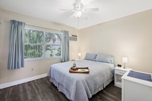 A bedroom with a bed, nightstands, a ceiling fan, a window with curtains, and a tray with a bottle and glasses. - Quaint Condo | Steps to the Beach and Dining (Marco Island)
