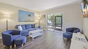 A modern living room with a blue sofa, a white coffee table, and a blue armchair. There is a painting of a beach scene on the wall, and a large window providing a view of the outdoors.