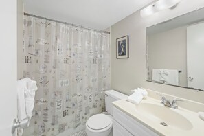A bathroom with a shower curtain featuring botanical prints, a white toilet, a sink with a mirror, and a towel rack. - Quaint Condo | Steps to the Beach and Dining (Marco Island)