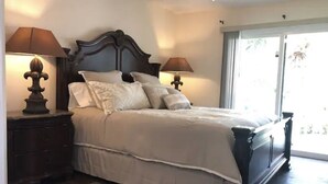Quarto com uma cama grande, duas mesas de cabeceira com luminárias, um painel de cabeceira de madeira e vista para o exterior através de uma porta de vidro deslizante.