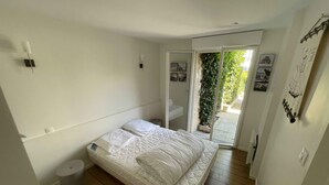 Un dormitorio con cama, un taburete pequeño, un inodoro blanco y un ventanal corredero que da a un balcón con vegetación.