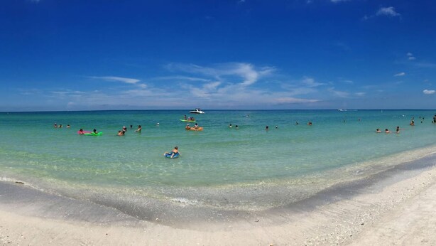 Una playa con agua turquesa cristalina, una orilla arenosa y algunos barcos a lo lejos.