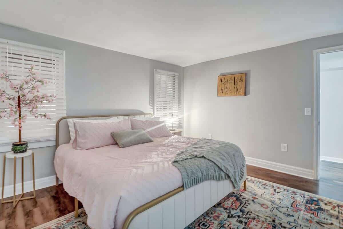 Un dormitorio con cama, ventana con persianas, una mesita con una planta y una pared decorada con una obra de arte.