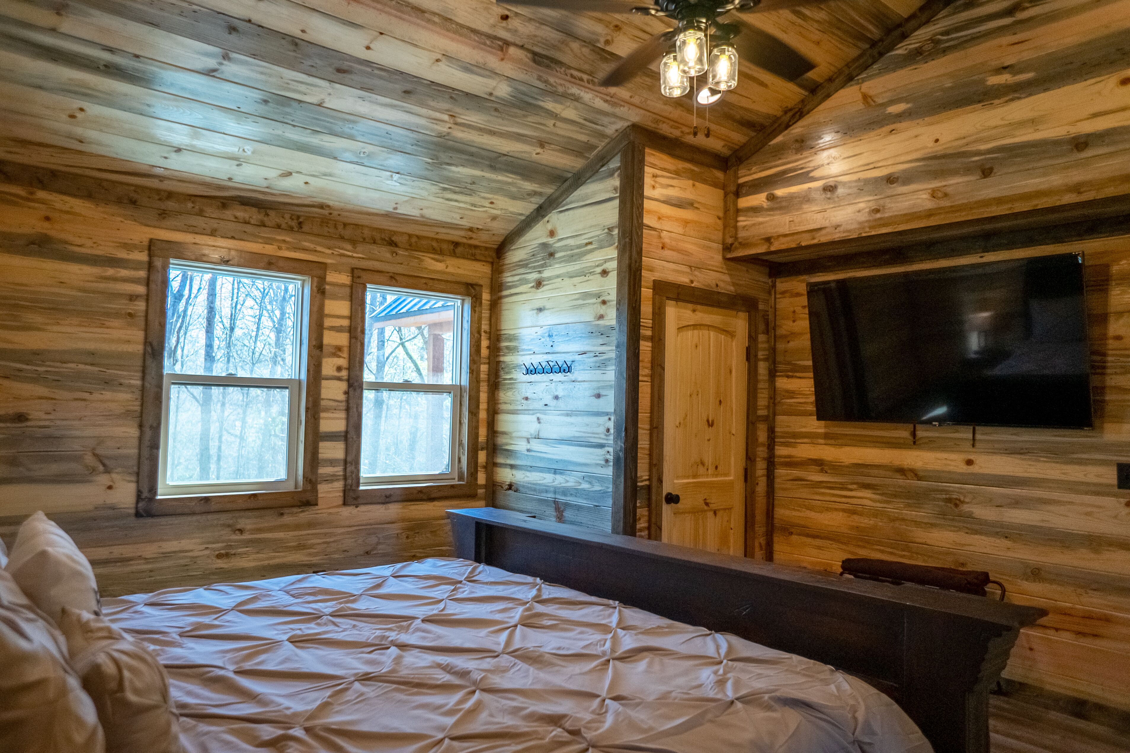 A wooden cabin room with a bed, a television, and two windows.