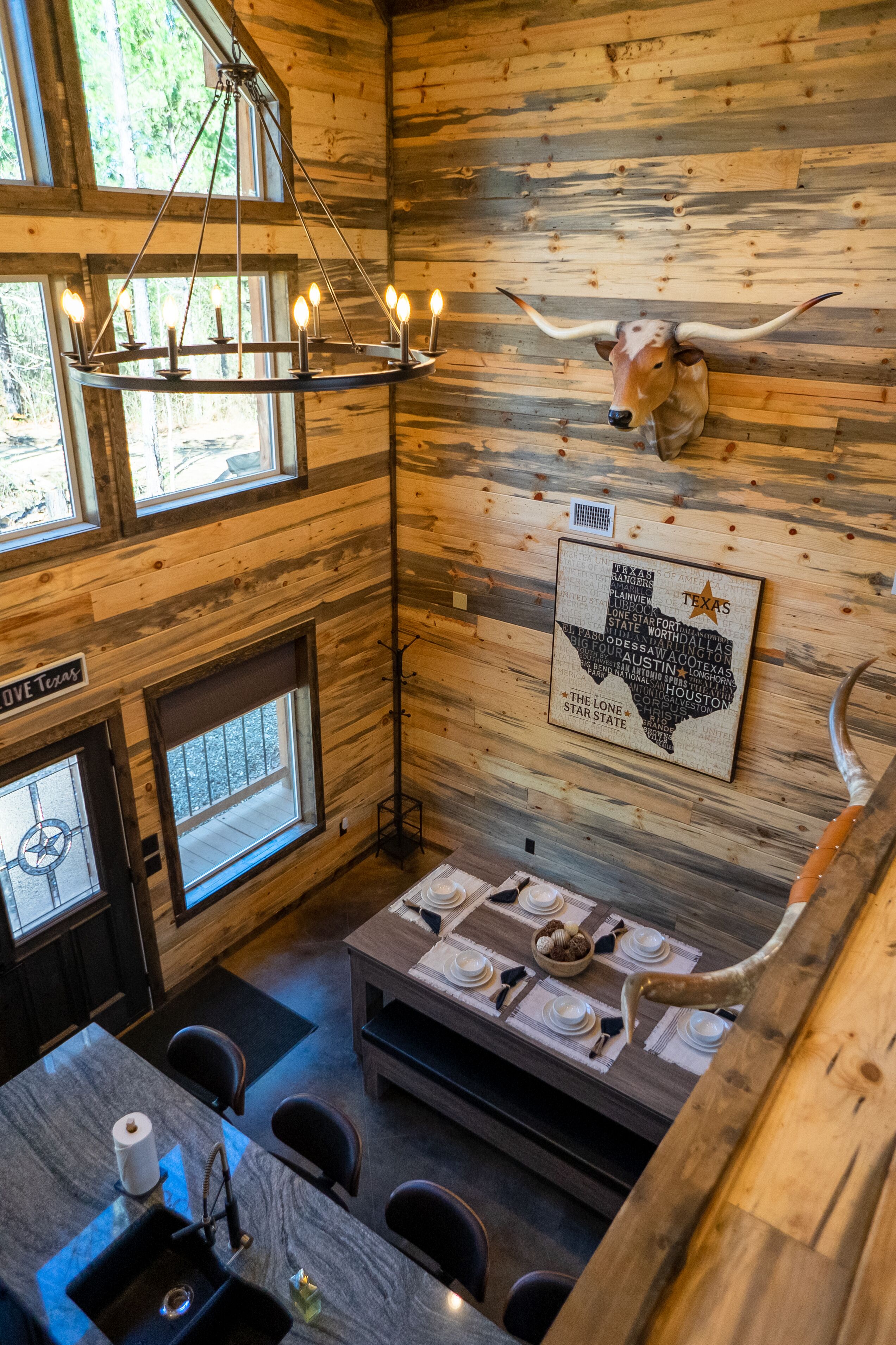 Un intérieur rustique avec des murs en bois, une tête de vache accrochée au mur, une décoration murale sur le thème du Texas, un lustre et un coin repas avec une table dressée pour quatre personnes.